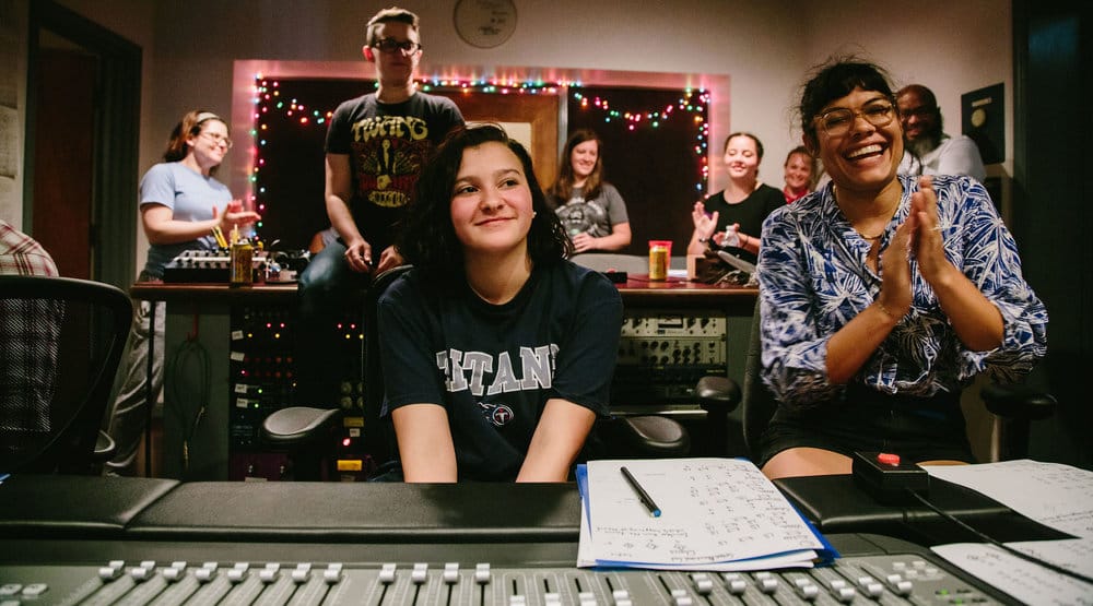 Young girl smiling in an audio recording studio with teacher clapping beside her